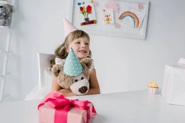Adorável aniversariante garoto sentado com ursinho de pelúcia à mesa com caixa de presente — Fotografia de Stock