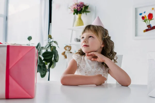 Niño de cumpleaños reflexivo mirando hacia otro lado y sentado en la mesa con el presente - foto de stock