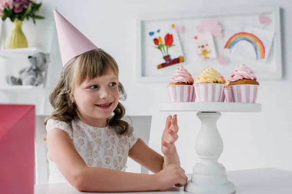 Niño de cumpleaños sonriente en cono apuntando a mano en el soporte con cupcakes - foto de stock