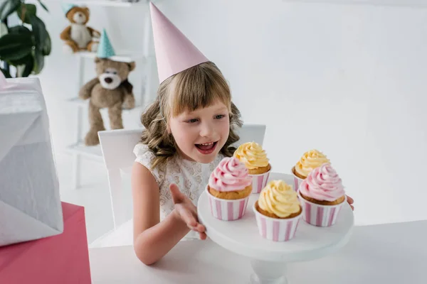 Niño de cumpleaños sonriente en cono sosteniendo soporte con cupcakes en la mesa - foto de stock