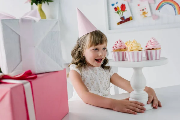 Aufgeregtes Geburtstagskind in Kegel schaut Cupcakes an, während es mit Geschenken am Tisch sitzt — Stockfoto