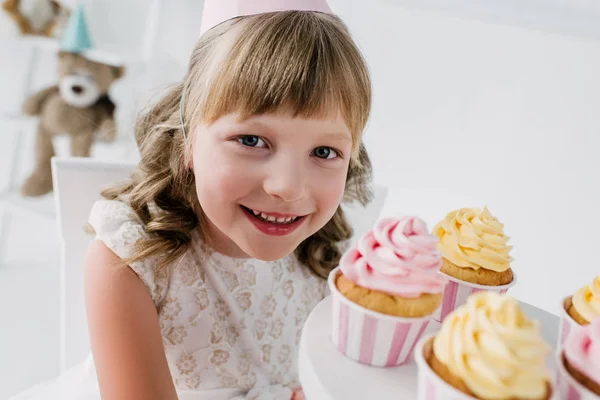 Close up portrait of smiling birthday kid in cone looking at camera and showing cupcakes — Stock Photo