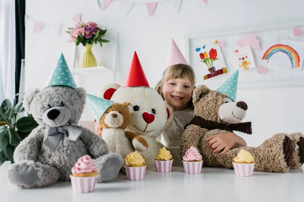 Sorrindo aniversário criança sentada com ursinhos de pelúcia em cones à mesa com cupcakes — Fotografia de Stock