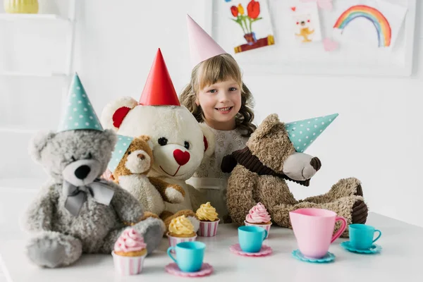 Enfant souriant anniversaire avec des ours en peluche dans des cônes ayant thé avec cupcakes à la table — Photo de stock