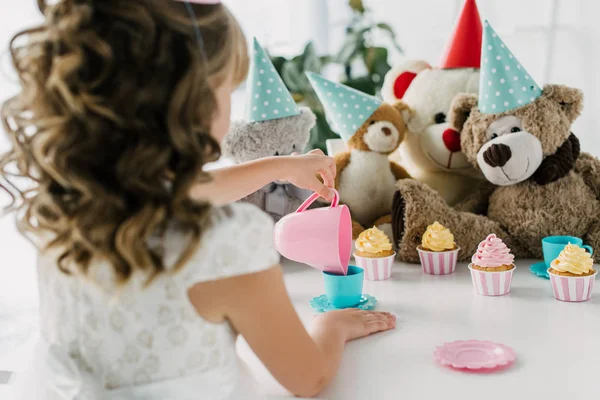 Vista trasera del niño de cumpleaños teniendo fiesta de té con osos de peluche en conos en la mesa con cupcakes - foto de stock