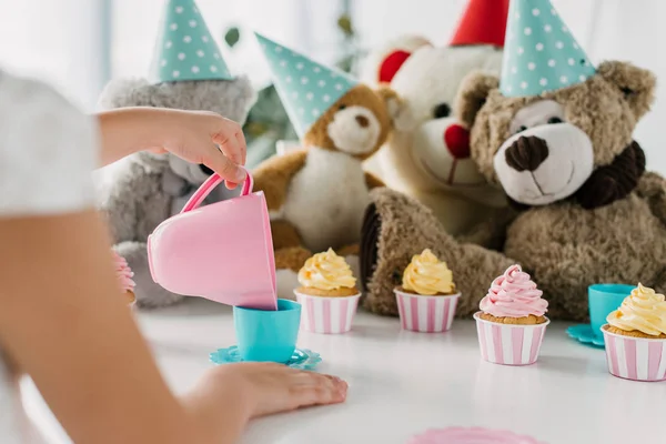 Imagem cortada de criança derramando chá em xícara para ursinhos de pelúcia em cones à mesa com cupcakes — Fotografia de Stock