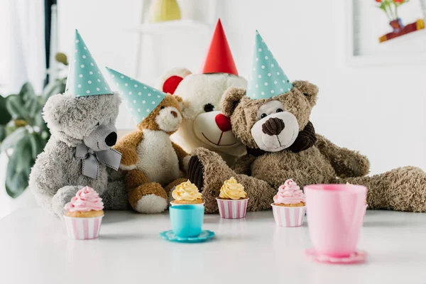 Teddy bears in cones on table with tea and cupcakes — Stock Photo