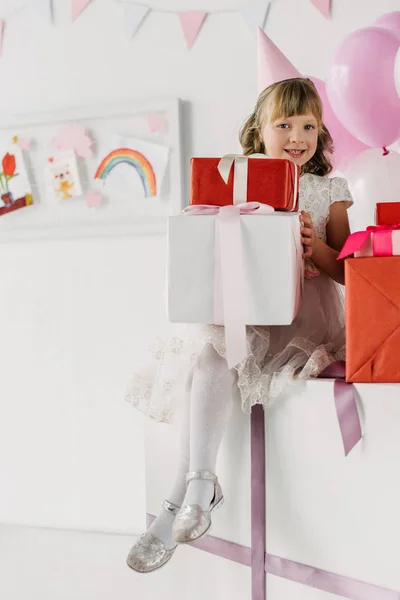 Souriant anniversaire fille dans cône tenant des boîtes-cadeaux et en regardant la caméra — Photo de stock