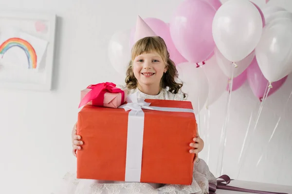 Petite fille d'anniversaire dans le cône tenant des boîtes-cadeaux et regardant la caméra — Photo de stock