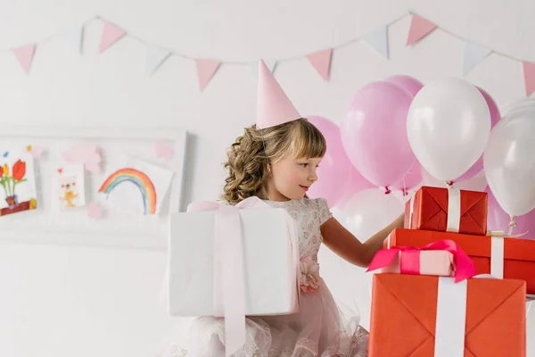 Carino compleanno bambino in cono guardando scatole regalo — Foto stock