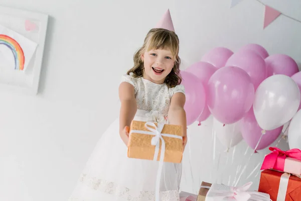 Щаслива дитина на день народження в конусі дивиться на камеру і показує подарункову коробку — стокове фото