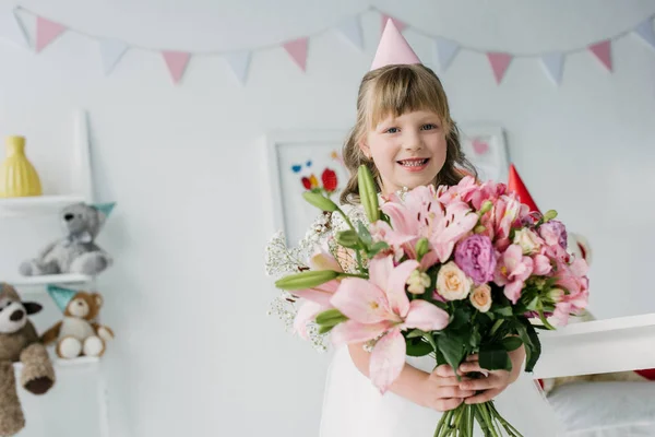 Happy birthday child in cone looking at camera and holding bouquet of lilies — Stock Photo