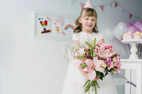 Smiling birthday child in cone looking at camera and holding bouquet of lilies — Stock Photo