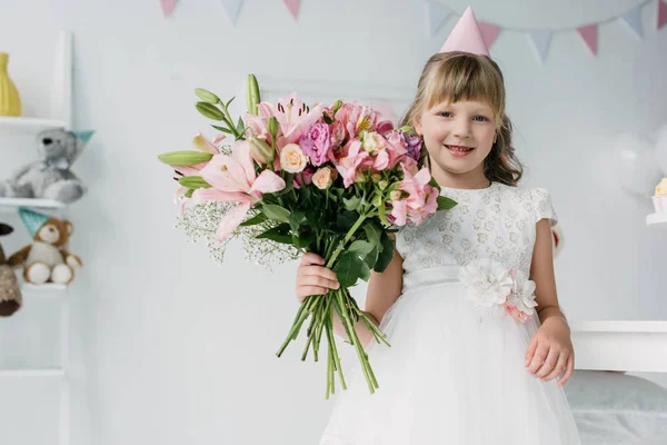 Smiling birthday kid in cone looking at camera and holding bouquet of lilies — Stock Photo