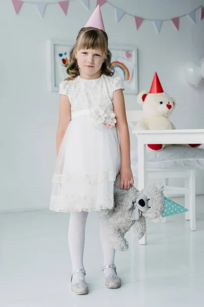 Pequena menina de aniversário ofendida em cone segurando ursinho de pelúcia — Fotografia de Stock