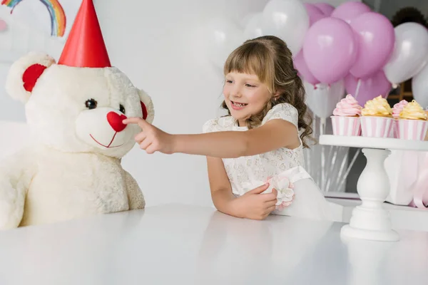 Smiling birthday kid touching nose of teddy bear in cone — Stock Photo