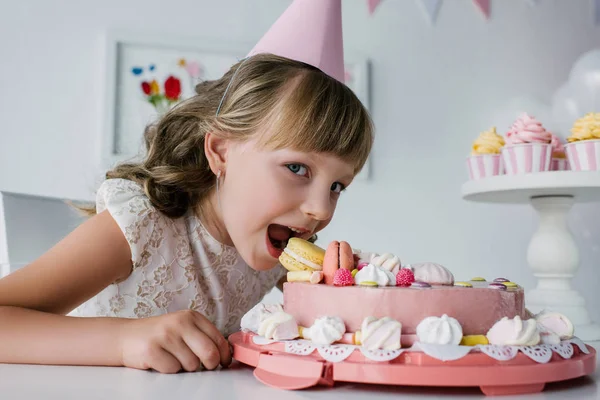 Lächelndes kleines Kind in Kegel, das Geburtstagstorte isst und in die Kamera schaut — Stockfoto