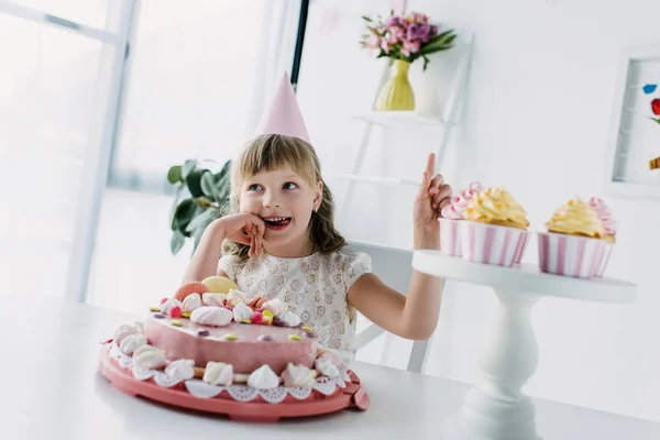Smiling birthday kid in cone doing idea gesture while sitting at table with cake — Stock Photo