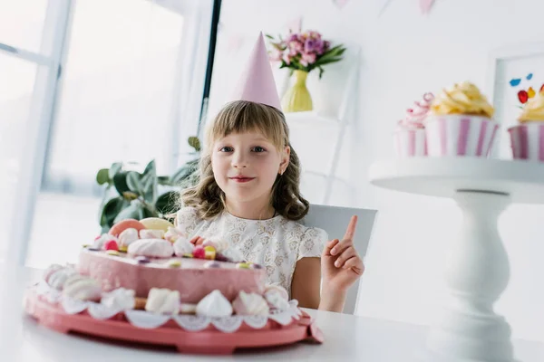 Happy child in cone doing idea gesture at table with birthday cake — Stock Photo
