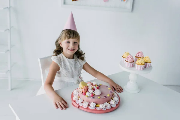 Vista ad alto angolo di adorabile bambino in cono seduto a tavola con torta di compleanno — Foto stock