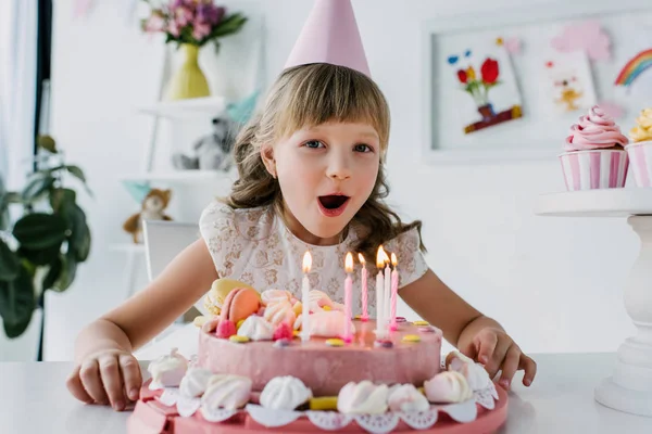 Smiling kid in cone blowing out candles from birthday cake — Stock Photo