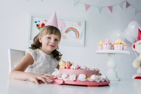 Happy adorable child in cone sitting at table with birthday cake — Stock Photo