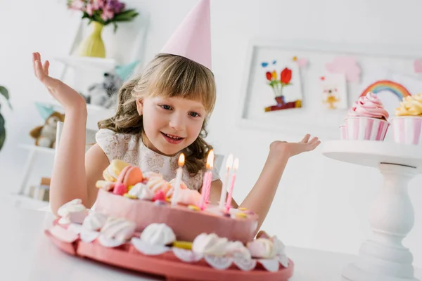 Улыбающийся маленький ребенок с широкими руками, смотрящий на праздничный торт со свечами — стоковое фото