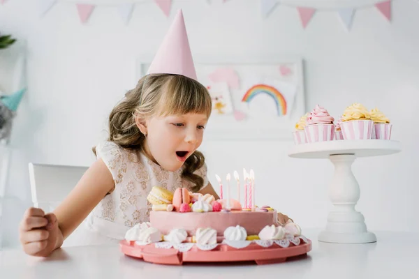 Little birthday girl in cone blowing out candles from cake on table — Stock Photo