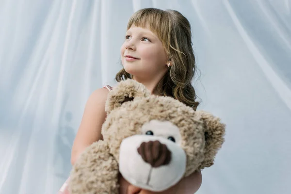 Sonriente niño pequeño mirando hacia otro lado y abrazando osito de peluche - foto de stock