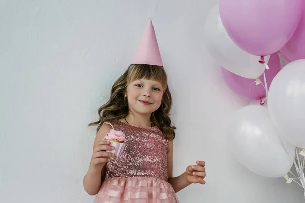 Happy birthday child in cone with dirty nose holding cupcake near pink balloons — Stock Photo