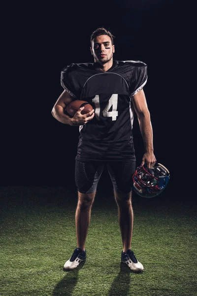 Confident american football player in black uniform holding helmet and ball and looking at camera on black — Stock Photo
