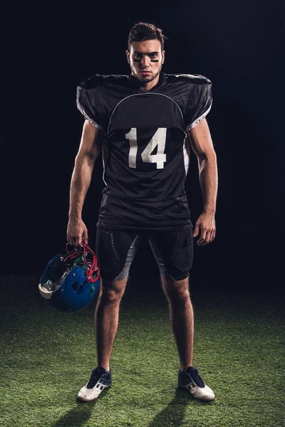 Confiante jogador de futebol americano em uniforme preto segurando capacete e olhando para a câmera em preto — Fotografia de Stock