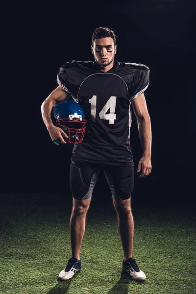 Serio giocatore di football americano in uniforme nera con casco e guardando la fotocamera su nero — Foto stock