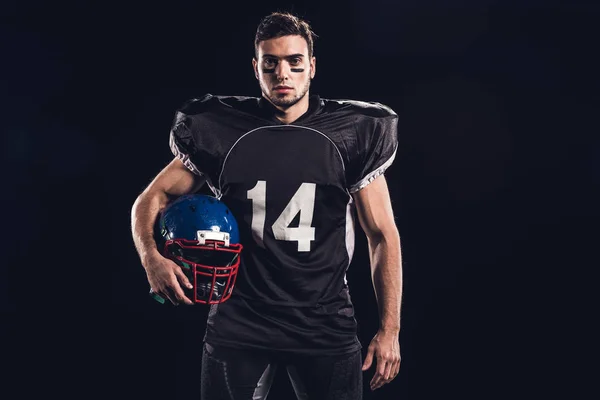 Joven futbolista americano en uniforme negro sosteniendo casco y mirando a la cámara aislada en negro - foto de stock