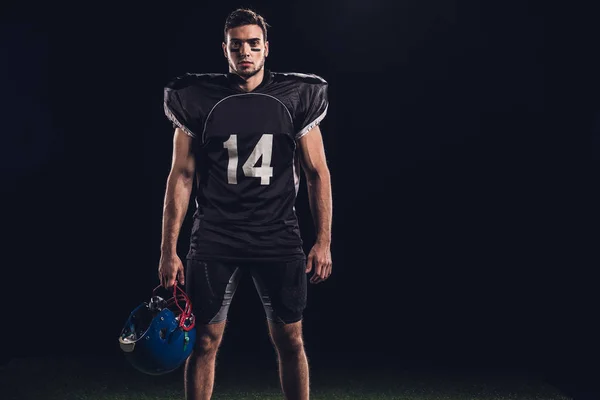 Guapo jugador de fútbol americano en uniforme negro sosteniendo casco y mirando a la cámara aislada en negro - foto de stock