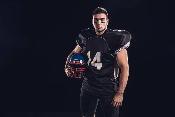 Atlético jugador de fútbol americano en uniforme negro con casco y mirando a la cámara aislada en negro - foto de stock