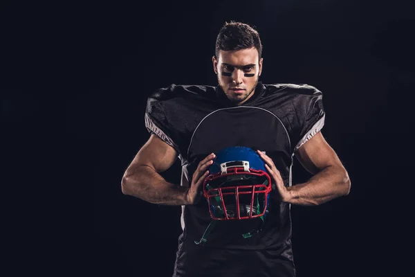 Jugador de fútbol americano sosteniendo el casco en las manos y mirando a la cámara aislada en negro - foto de stock