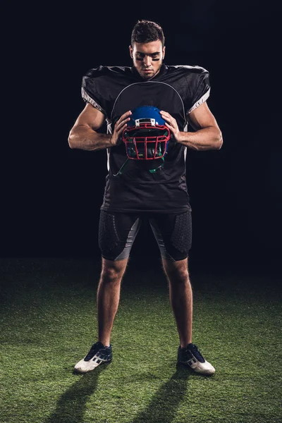 Jogador de futebol americano em pé na grama verde e segurando capacete em mãos em preto — Fotografia de Stock