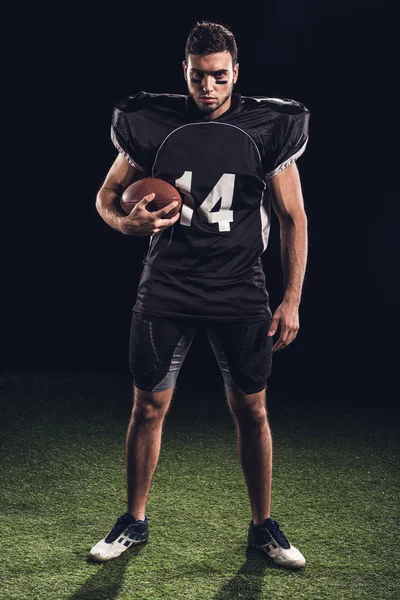 Junger selbstbewusster amerikanischer Fußballspieler mit Ball in der Hand, der in die Kamera schaut, während er auf schwarzem Gras steht — Stockfoto