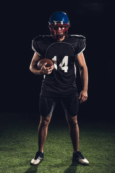 Confident american football player holding ball in hand and looking at camera isolated on black — Stock Photo