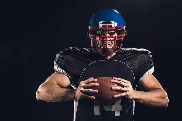 Verrückter amerikanischer Fußballspieler hält Ball in den Händen und schaut in die Kamera isoliert auf schwarz — Stockfoto