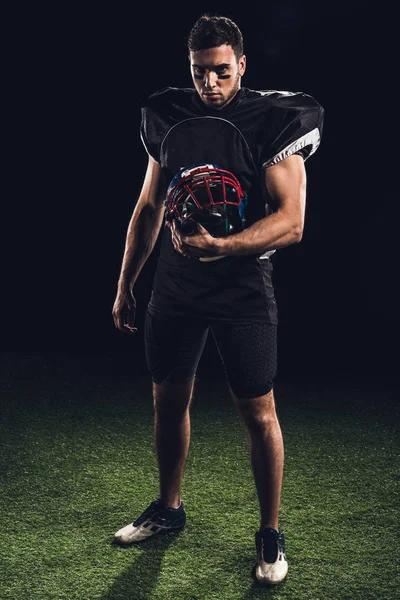 Jugador de fútbol americano serio en uniforme negro con casco y mirando a la cámara en negro - foto de stock