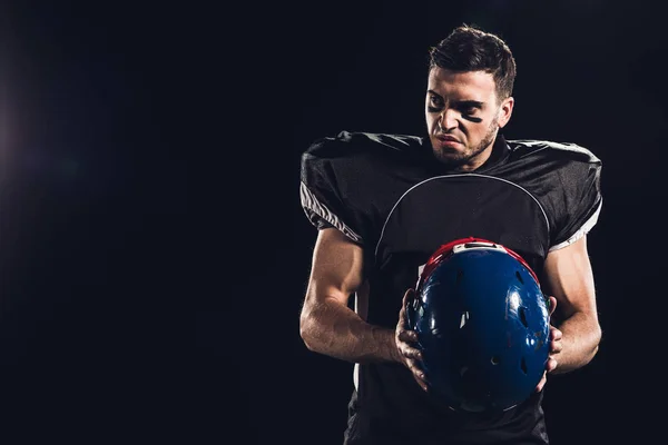 Jugador de fútbol americano enojado en uniforme negro con casco y mirando hacia otro lado aislado en negro - foto de stock