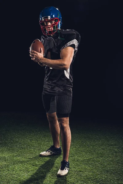 American football player standing on grass with ball on black and looking at camera — Stock Photo