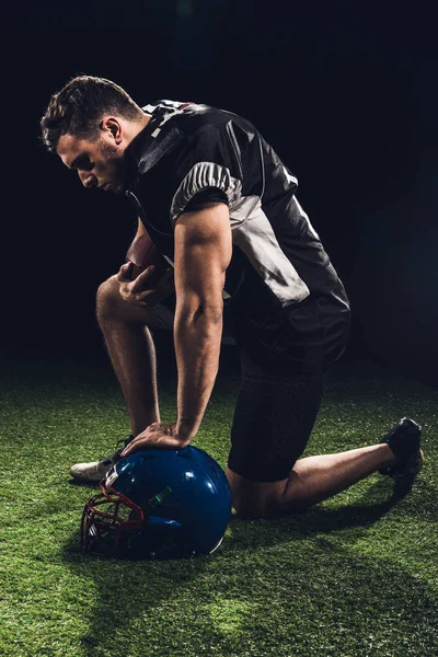 Joven jugador de fútbol americano serio de pie sobre una rodilla en la hierba con casco en negro - foto de stock