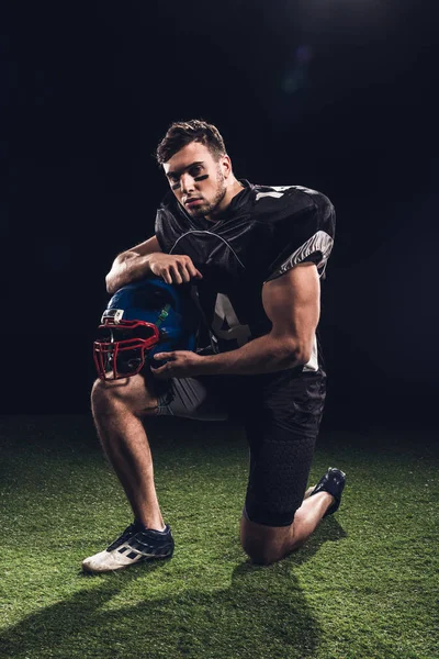 Guapo jugador de fútbol americano de pie sobre una rodilla en la hierba con casco en negro - foto de stock