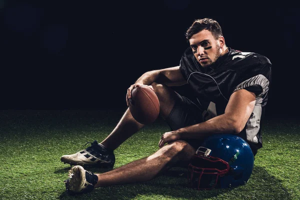 Американський футболіст, сидячи на траві з м'ячем і шолом, на чорному — стокове фото