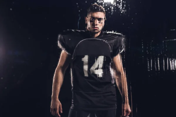 Перегляд молодий американський футболіст дивлячись на камеру на чорному через мокрій скло — стокове фото