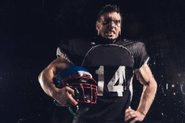 Vue du joueur de football américain en colère avec casque sur noir à travers verre mouillé — Photo de stock