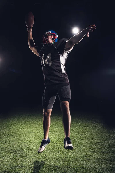 Athlète américain joueur de football sautant avec balle sous les projecteurs sur noir — Photo de stock
